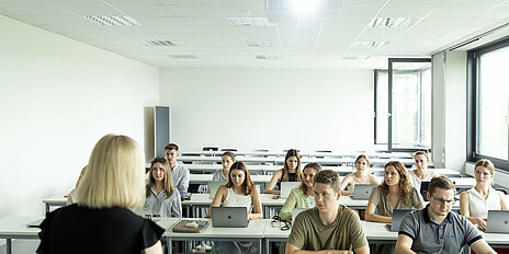 Eine Gruppe von 13 Personen sitzt in einem Hörsaal. Die Fenster sind geöffnet.