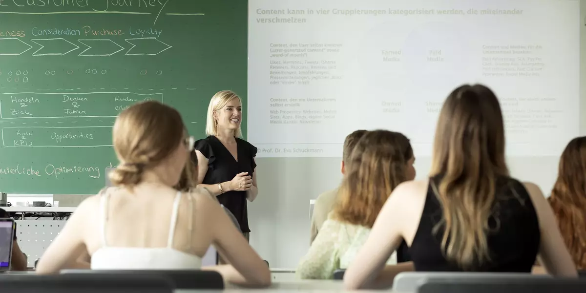 Studierende sitzen in der Vorlesung bei Prof. Dr. Schuckmann. Links im Bild ist eine grüne Tafel.