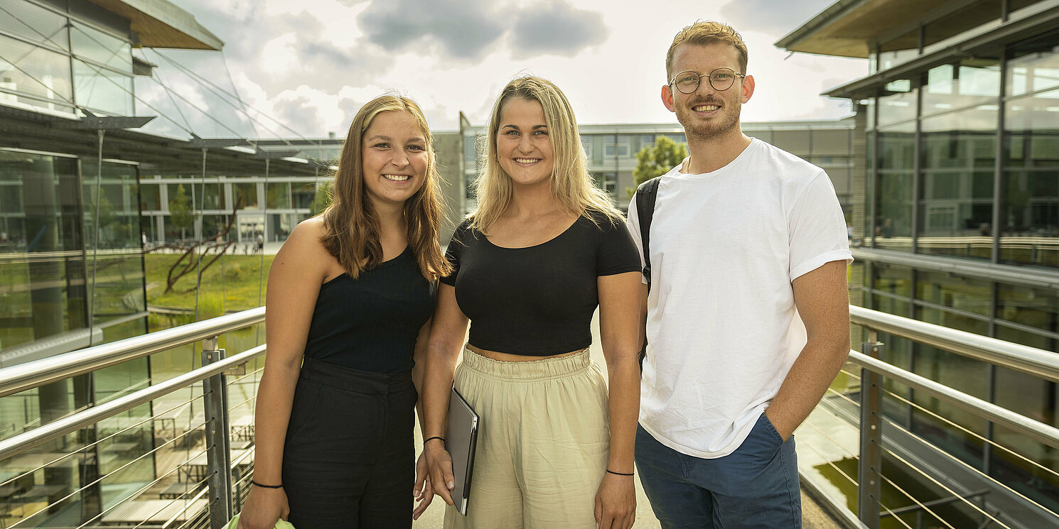 Zwei Frauen und ein Mann stehen auf der OTH Brücke. Rechts davon die Mensa, links die Bibliothek.