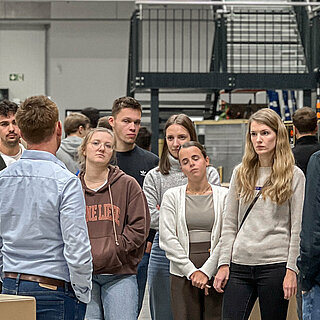 Eine Gruppe von Studierenden stehen in einem Raum, im Hintergrund Pakete und Regale