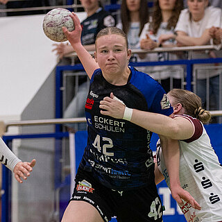 Wettkampfszene beim Handballspiel in der Halle: Milena Stürenburg in der Mitte, in begriff den Ball zu werden, wird umrahmt von zwei gegnerischen Spielerinnen. Im Hintergrund Zuschauer auf der Tribüne.